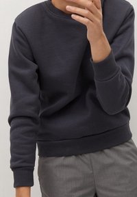 Tmavě šedá mikina s kulatým výstřihem, dlouhými rukávy a žebrovanými manžetami. Hladká textura a volný střih, kombinováno se šedými kalhotami.