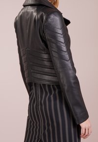 Mustasuinen nahkainen lyhyt takki, jossa on vaakasuorat ja chevron-muotoiset uritukset selässä ja hartioissa, sisältäen klassisen kauluksen ja vetoketjusulkemisen.