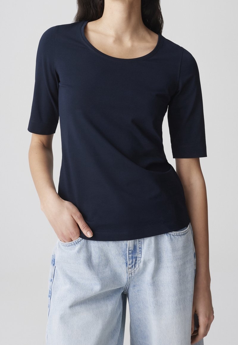 Femme portant un haut bleu marine à manches courtes et un jean denim bleu clair, une main dans la poche, devant un fond uni.