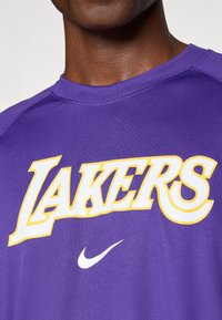 T-shirt de sport violet en tissu texturé, arborant "LAKERS" en lettres blanches et jaunes en gras. Comprend un petit logo Nike blanc.