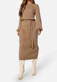 Femme portant une robe pull côtelée camel avec de longues manches bouffantes, une ceinture assortie et des bottes crème montantes jusqu'aux genoux sur fond blanc.