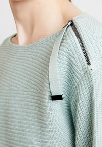 Himmelblauer, strukturierter Pullover mit rundem Ausschnitt, geripptem Muster und einem Reißverschlussdetail auf einer Schulter mit einem Stoffband und schwarzen Akzenten.
