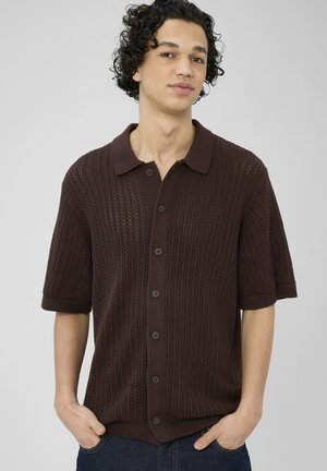 Jeune homme aux cheveux bouclés portant une chemise tricotée marron foncé à manches courtes avec boutons, mains dans les poches, fond gris neutre.