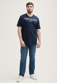 Camiseta de algodón azul marino con mangas cortas y cuello redondo, que presenta un texto de logo impreso en blanco. Combinada con jeans azules y zapatillas blancas.