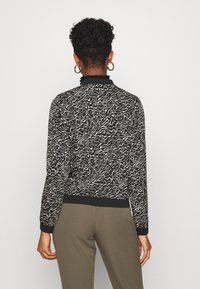 Žena s krátkými kudrnatými vlasy, která má na sobě zlaté kruhové náušnice, černý a béžový top s abstraktním vzorem a olivové kalhoty, otočená zády.