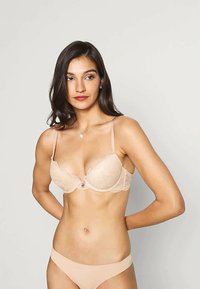 Žena s dlouhými tmavými vlasy, oblékající béžovou krajkovou podprsenku a odpovídající kalhotky, stojící s rukama za zády na jednobarevném světlém pozadí.