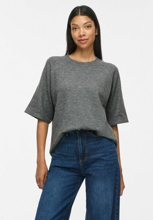 Kvinde iført en løs grå kortærmet trøje og højtsiddende blå jeans, stående med hænderne afslappet ned langs siderne.