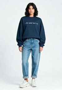 Mornarskoplavi oversized dukserica s natpisom, uparena s svijetloplavim trapericama s prevrnutim rubovima i bijelim tenisicama. Tkanina je mekana, ležeran dizajn.