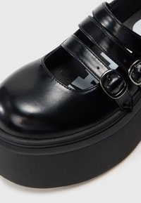 Zwarte leren schoen met ronde neus, voorzien van twee verstelbare riemen en gespen. Dikke, gestructureerde zwarte platformzool.
