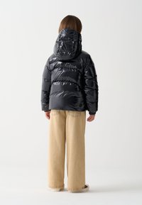 Melnā pūtuma jaka ar kapuci, spīdīgu virsmu, kvadrātveida šuvumu rakstu un elastīgām aprocēm, kopā ar bēša krāsas platām biksēm uz neitrāla fona.