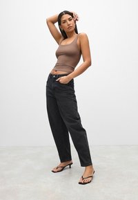 Pruun sobiv tank top, kõrge vöökohaga mustad teksad ja mustad kontsaga rihmaflipflopid. Mudel poseerib, üks käsi puusal ja teine käsi üles tõstetud.