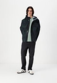 Dark green waterproof jacket with hood, front zip, and two large chest pockets. Worn over a light green shirt and black jeans with sneakers.
