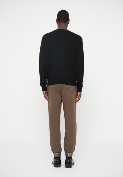 Man standing facing away wearing a black sweater, brown jogger pants with a back pocket, and black dress shoes.