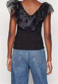 Bakifrån av en person som bär en svart volanglös ärmlös topp med genomskinligt tyg vid axlarna och blå högmidjade jeans.
