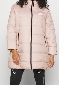 Person som bär en ljusrosa Nike-puffjacka med svart dragkedja och sidofickor, i kombination med svarta leggings som har vita Nike-loggor.