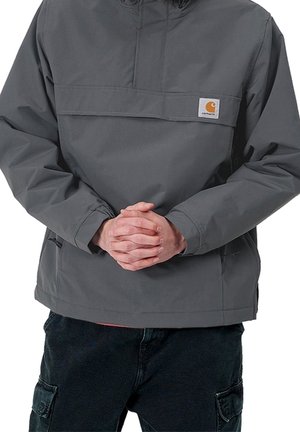 Person iført en grå Carhartt-jakke med foldede hænder hvilende ved livet, kombineret med sorte cargobukser.