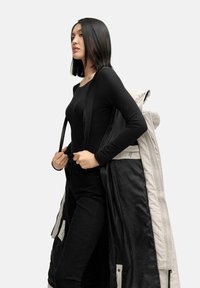 Zwarte lange mouw top, zwarte broek en een beige pufferjack met een zwarte voering. Het jack is open en over de arm gehangen.