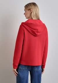 Felpa rossa con tessuto strutturato, dotata di cappuccio e spalle scese, abbinata a jeans blu. Tasche posteriori visibili sui jeans.