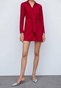 Robe chemise rouge avec un détail noué à l'avant, manches longues et col. Associée à des talons argentés à bout pointu avec des brides à la cheville. Texture lisse.