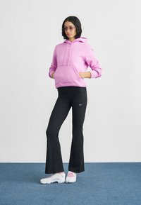 Ung kvinne iført rosa hettegenser, svarte utsvingte leggings med Nike-logo, hvite joggesko og tonede briller står med hendene i lommene.