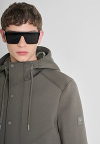 Grau kapuzenjacke mit glatter Textur, ausgestattet mit Knöpfen und Zugbändern. Getragen mit kantigen schwarzen Sonnenbrillen, die ein zeitgemäßes Design betonen.