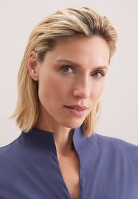 Blauwe blouse met een hoge kraag, gladde textuur, aansluitend model en subtiele glans. Model met lichtblond haar en neutrale make-up kijkt recht in de camera.