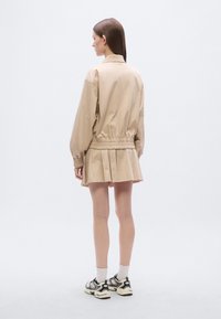 Μπεζ, ελαφρύ μπουφάν με σούρα στη μέση, συνδυασμένο με πλισέ μίνι φούστα. Το μοντέλο φοράει μαύρα και άσπρα αθλητικά παπούτσια με καλτσάκια μέχρι τον αστράγαλο.