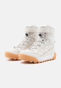 Botas de senderismo blancas con un upper texturizado, correa ajustable y suela de goma naranja. Incluye ganchos y acentos reflectantes para mayor visibilidad.
