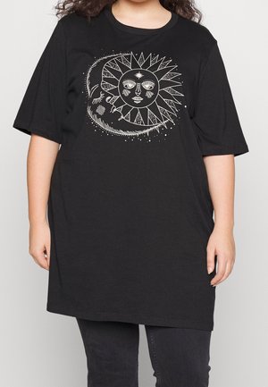 Person trägt ein schwarzes Oversize-T-Shirt mit einer weißen Grafik eines Sonnen- und Sichelmondgesichts auf der Vorderseite, kombiniert mit dunkler Hose.