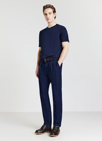 Giovane uomo in piedi con le mani in tasca, indossa una maglietta blu navy, jeans blu scuro, cintura marrone e scarpe lacciate marrone scuro su sfondo bianco.