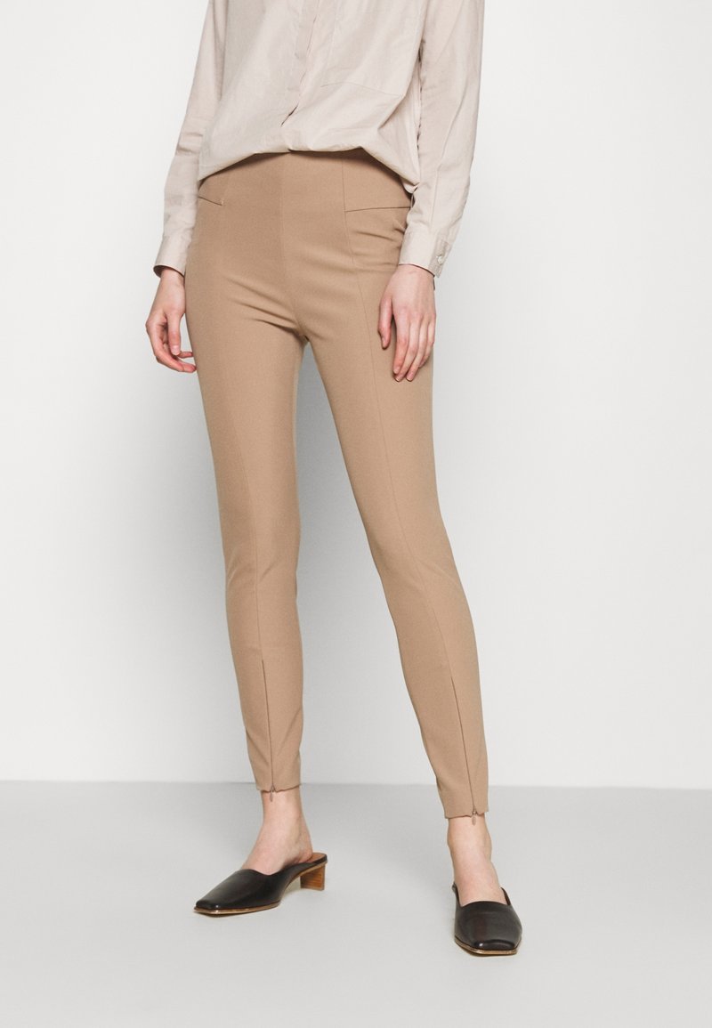 Pantalon beige coupe slim en tissu texturé, avec un devant plat et des poches latérales. Associé à une chemise beige clair et des mules noires.