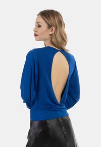Blaues langärmliges Oberteil mit tiefem Ausschnitt im Rücken, mit einem Wickeldetail an der Taille. Glatte Stoffqualität mit weicher Textur, kombiniert mit einem schwarzen Rock.