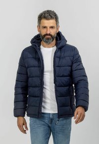 Tamsiai mėlynas pūkinis švarkas su gobtuvu, turintis siuvinėtas dalis, kišenes su užtrauktuku ir lygų paviršių. Dėvimas ant baltos marškinių.