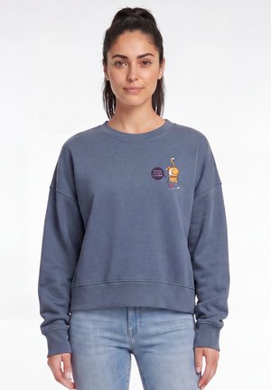 Femme aux cheveux foncés attachés en queue de cheval, portant un sweat-shirt bleu avec un petit chat cartoon et une bulle de dialogue sur la poitrine, assorti à un jean bleu clair.