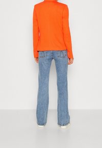 Blazer cintré orange avec col à revers, associé à un jean évasé bleu clair avec détail de surpiqûres sur les poches arrière. Texture du tissu lisse.