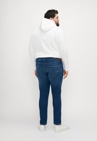Felpa bianca con cappuccio e jeans blu a gamba dritta. I jeans presentano tasche posteriori e una toppa in pelle marrone sulla vita. Sneakers bianche.