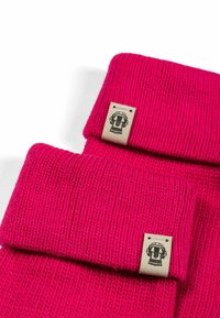 Zwei gefaltete, leuchtend pinke gestrickte Mützen mit beigen Anhängern, die schwarze dekorierte Logos auf weißem Hintergrund zeigen.