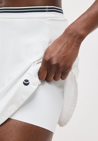 Ruka drží boky bielej športovej sukne s čiernym a zeleným pruhovaným pásom, odhaľujúc biele šortky pod ňou.