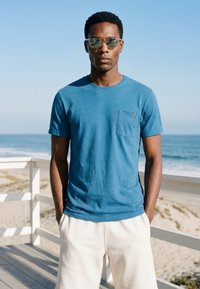 Young man wearing sunglasses, blue T-shirt, and white shorts stands on a wooden beach boardwalk with ocean waves in the background.