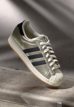 Metallisk sølv Adidas sneaker med sorte striber, hvide snørebånd og en gummiskal tå, der hviler på en struktureret stens overflade.