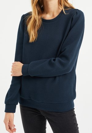 Femme portant un pull texturé bleu marine à manches longues avec un col rond et un pantalon noir, debout devant un fond blanc.