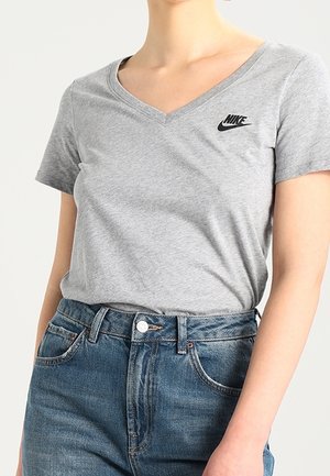 Kobieta nosząca jasnoszarą koszulkę Nike z dekoltem w serek, wpuszczoną w wysokie niebieskie dżinsy z kieszeniami z przodu i szlufkami.
