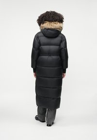 Zwarte pufferjas met een met bont gevoerde capuchon, met blokachtige segmenten en een lange lengte, gecombineerd met losse grijze spijkerbroek en zwarte schoenen.