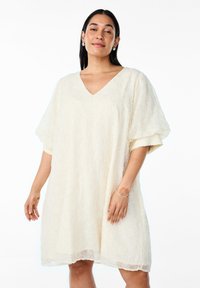 Robe courte couleur crème à manches courtes avec un col en V, motif floral texturé, coupe décontractée et ourlet semi-transparent.