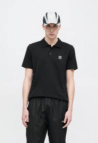 Mustast polo särgist, millel on kolme nupuga lõhik ja väike valge Adidas logo. Kandmisel on seljas mustad, lahtised püksid. Nokamütsil on hallid ja mustad triibud.