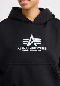 Čierna mikina s kapucňou vyrobená z jemnej bavlny s prednou vreckom. Obsahuje biele potlačené logo a text: "ALPHA INDUSTRIES, KNOXVILLE, TENNESSEE, U.S.A."