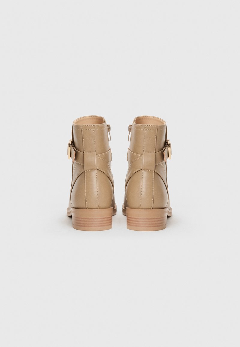 Botas de cuero beige a los tobillos con punta redonda, cremallera lateral y un tacón bajo apilado. Presenta costuras entrecruzadas y una hebilla decorativa.