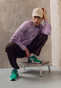 Camisa de manga larga morada, pantalones marrones, zapatillas verdes con rayas blancas y una gorra beige. Modelo agachado en un banco metálico.