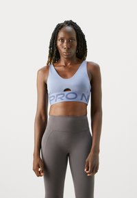 Mujer atlética con cabello trenzado está de frente, vistiendo un sujetador deportivo azul claro y leggings grises de cintura alta, frente a un fondo simple.