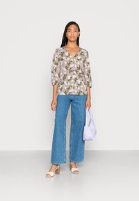 Blusa floral em verde-azeitona com flores roxas e brancas, combinada com jeans ampla azul, sandálias brancas de salto bloco e uma mala lavanda.